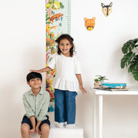 Two children standing next to a colorful growth chart with animal illustrations on a white wall.