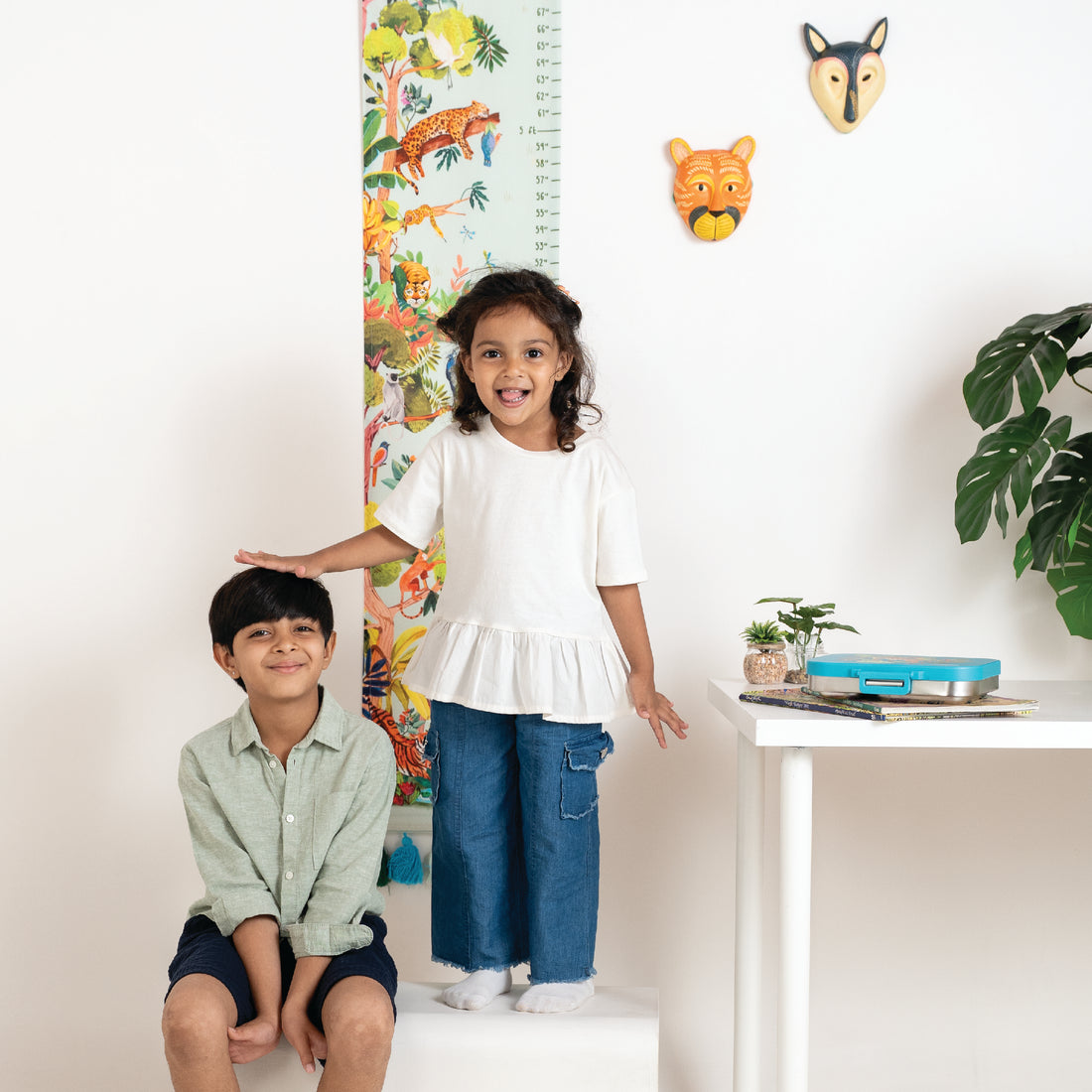 Two children standing next to a colorful growth chart with animal illustrations on a white wall.