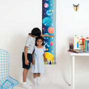 Two children using a colorful height chart in a room with a table and chair.