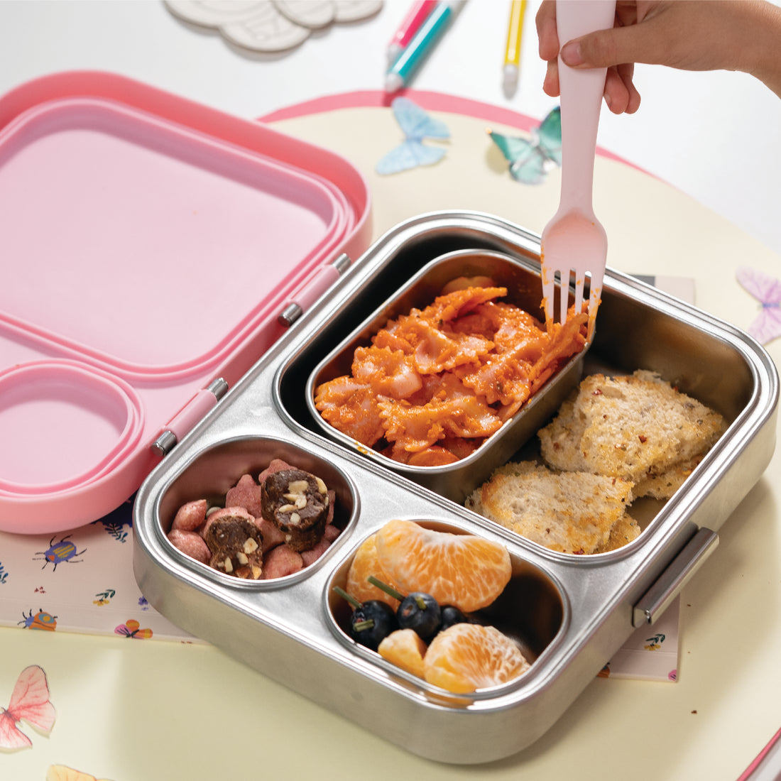 Metal bento box with compartments of food, pink lid, and white fork on a colorful background.
